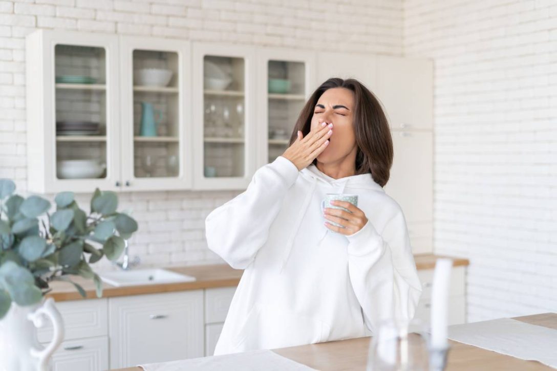 Young woman in a warm white hoodie at home in the kitchen, starts her day with a cup of coffee, yawns, barely woke up, lack of sleep