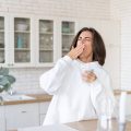 Young woman in a warm white hoodie at home in the kitchen, starts her day with a cup of coffee, yawns, barely woke up, lack of sleep