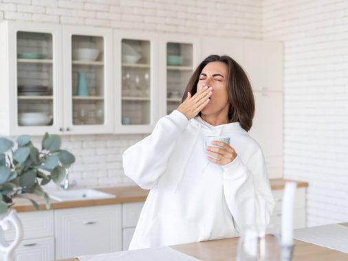 Young woman in a warm white hoodie at home in the kitchen, starts her day with a cup of coffee, yawns, barely woke up, lack of sleep